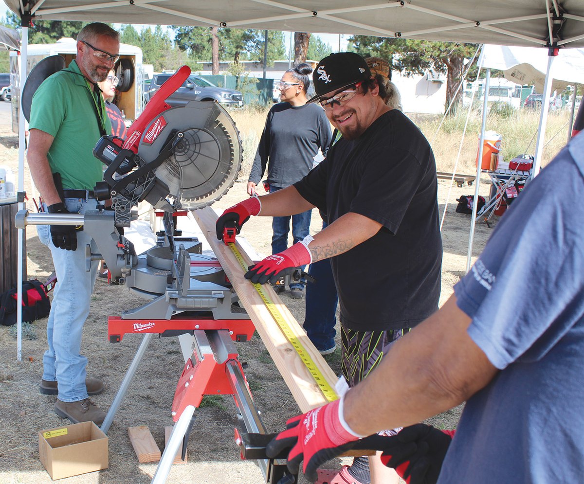 An instructor watches as a student measures a plank at a miter saw station under a canopy