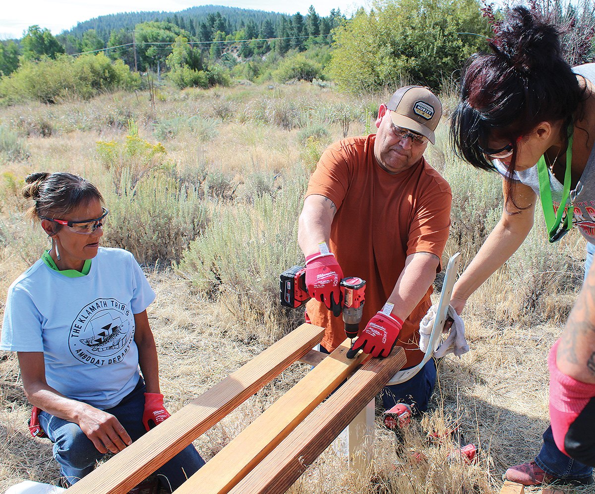 Three workers drill deck boards onto a park bench frame in a field
