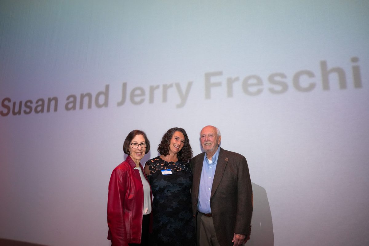 Susan and Jerry Freschi smiling with a guest on the Ross Ragland Theater stage, their names projected on the screen behind them