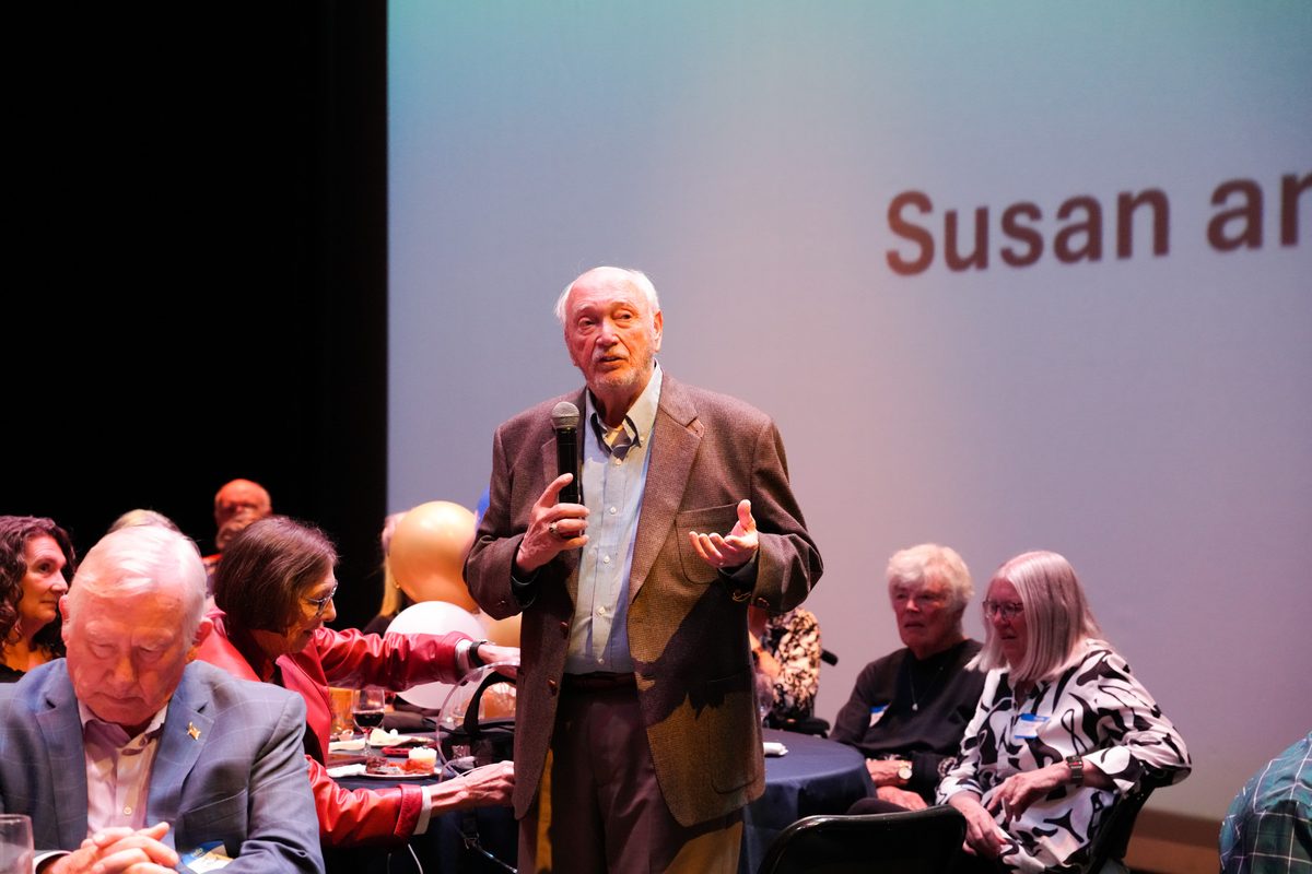 Jerry Freschi speaking into a microphone in front of the projected Susan and Jerry Freschi title screen