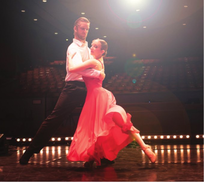 A pair of ballroom dancers mid-step on the Ross Ragland Theater stage, the dancer in a flowing red gown