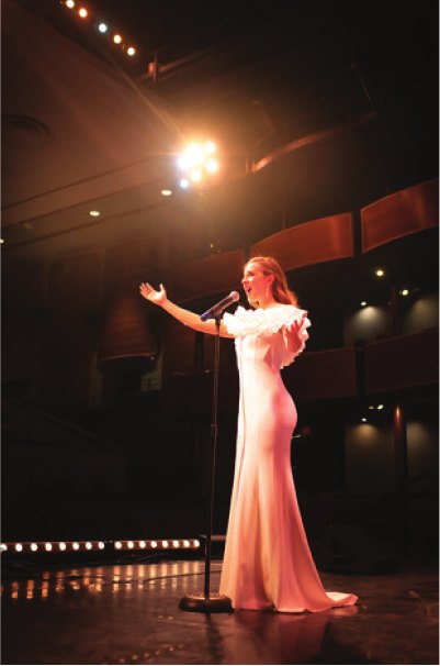 A soloist in a pale gown sings into a microphone on the Ross Ragland Theater stage