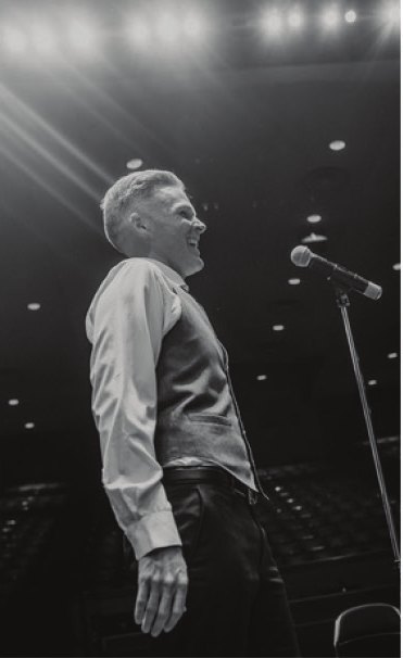 Black-and-white photo of a vocalist singing at a microphone stand under a stage spotlight