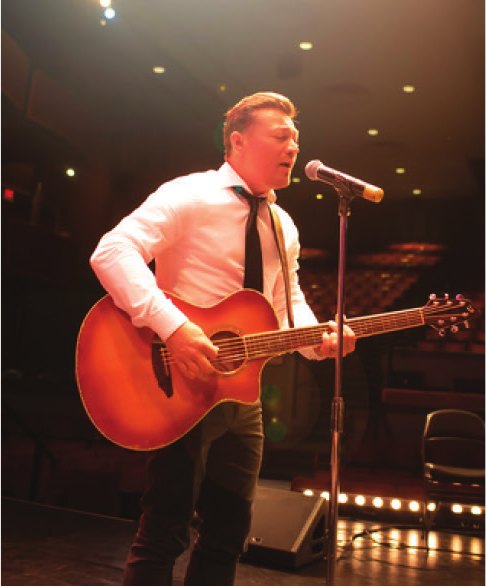 A guitarist in a white shirt and tie performs at the microphone on stage