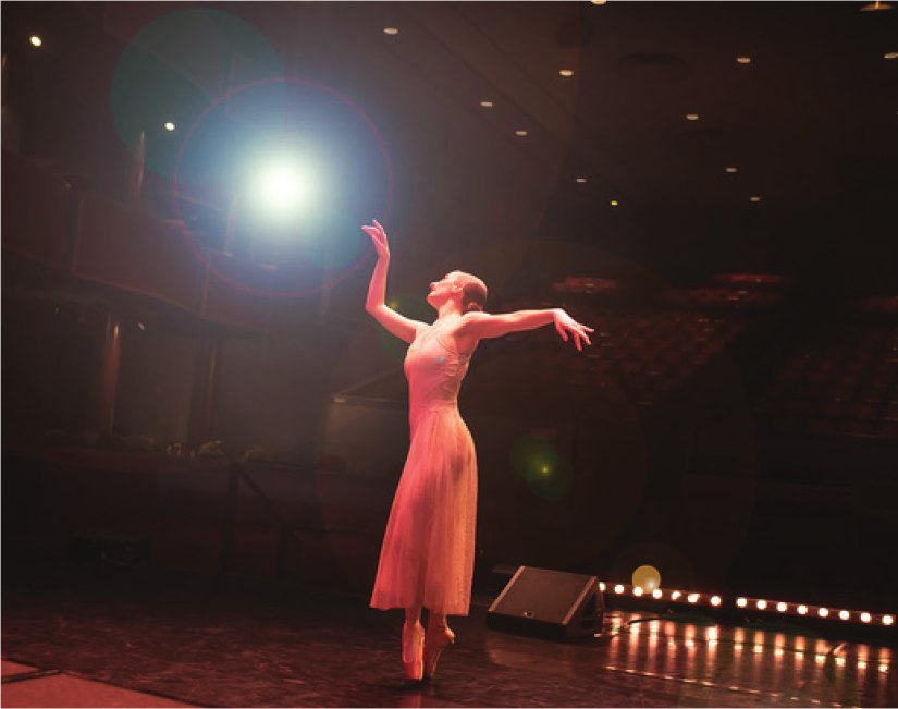 A ballerina on pointe reaches upward in a spotlight on the Ross Ragland Theater stage