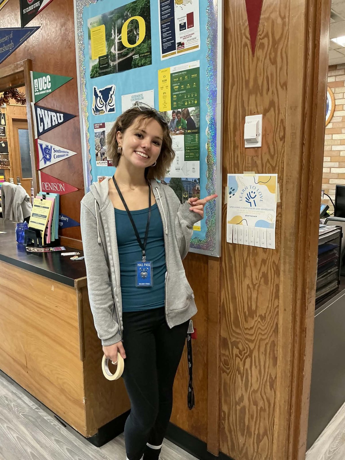 A student points to a Learn to Love card with take-one tabs posted beside a school office window