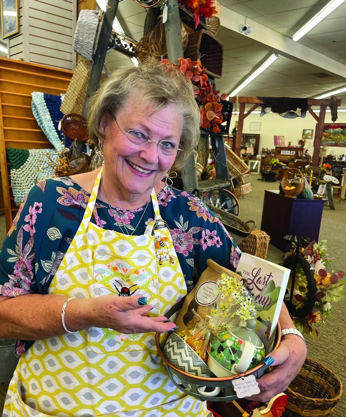 Smiling woman in a yellow apron holding a gift basket inside Honey & Ivy Collective in Klamath Falls
